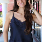 A woman with long brown hair smiling while wearing a navy blue satin camisole top with lace trim, standing in an outdoor walkway.