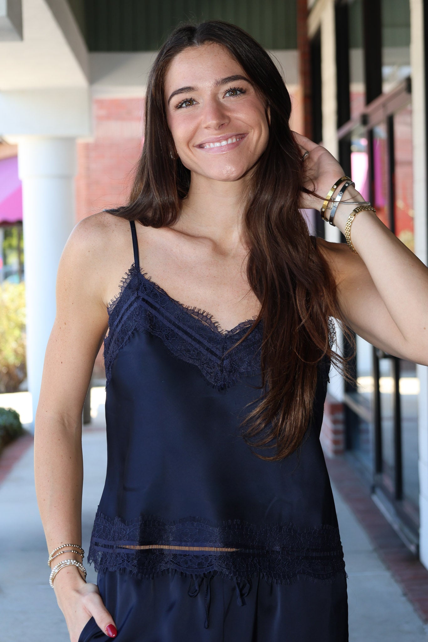 A woman with long brown hair smiling while wearing a navy blue satin camisole top with lace trim, standing in an outdoor walkway.