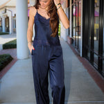 A woman wearing a navy blue satin jumpsuit with lace trim and wide legs, walking on an outdoor shopping center walkway.