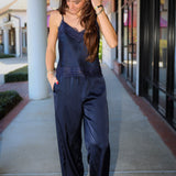 A woman wearing a navy blue satin jumpsuit with lace trim and wide legs, walking on an outdoor shopping center walkway.