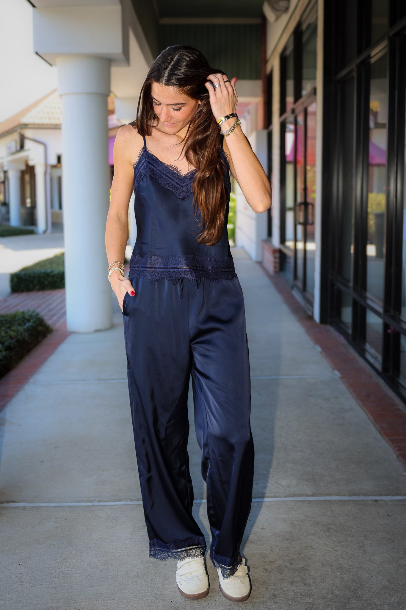 A woman wearing a navy blue satin jumpsuit with lace trim and wide legs, walking on an outdoor shopping center walkway.