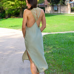 A woman wearing a sage green satin slip dress with an open back and high side slit, photographed outdoors on a paved path next to a grassy lawn.