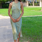 A woman wearing a sleeveless sage green midi dress with a side slit and white strappy heels, walking on a paved path in a park with green grass and trees.