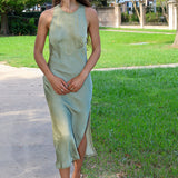 A woman wearing a sleeveless sage green midi dress with a side slit and white strappy heels, walking on a paved path in a park with green grass and trees.
