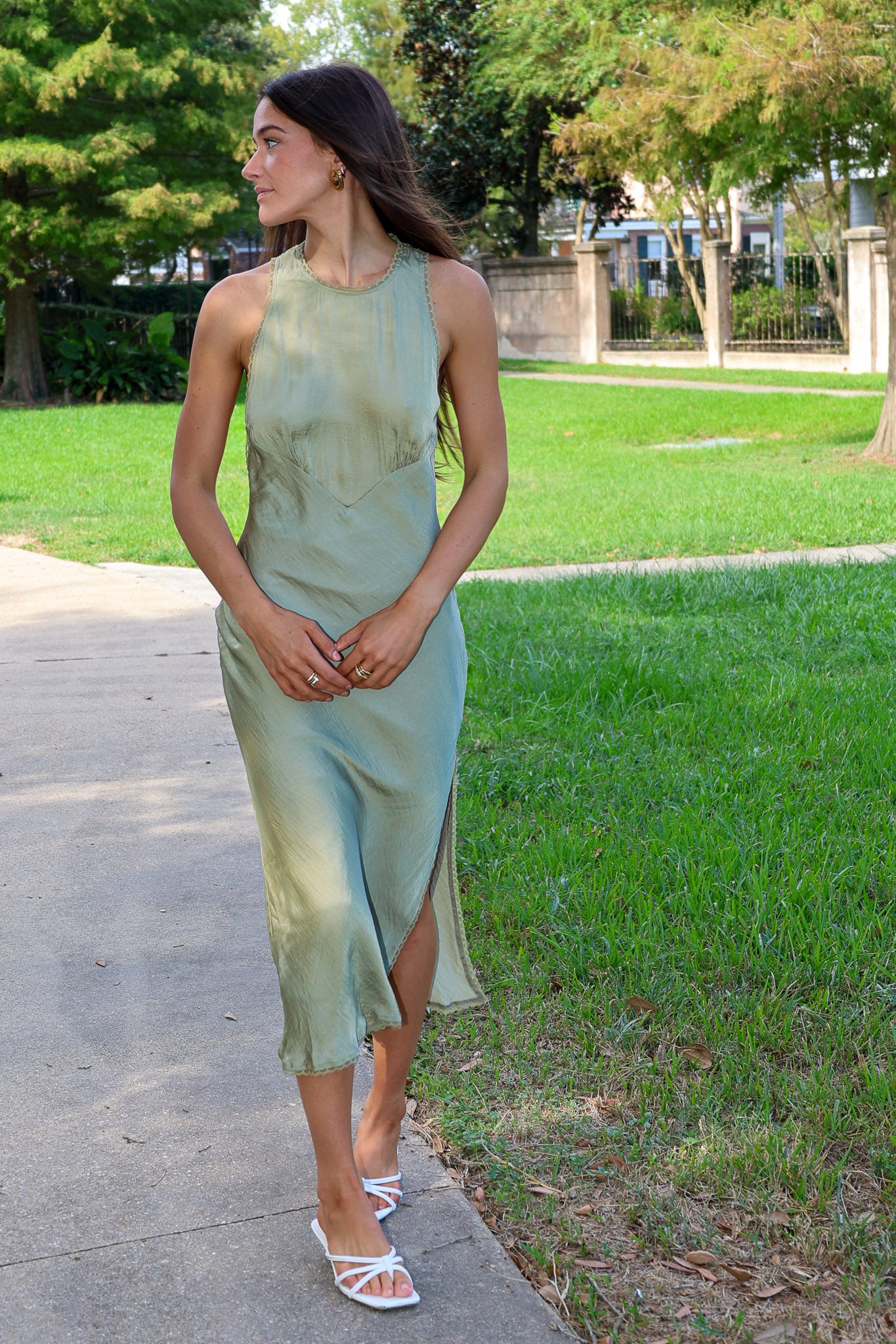 A woman wearing a sleeveless sage green midi dress with a side slit and white strappy heels, walking on a paved path in a park with green grass and trees.