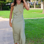 A woman wearing a sleeveless olive green midi dress and white strappy sandals walking on a paved path in a park with green grass and trees.