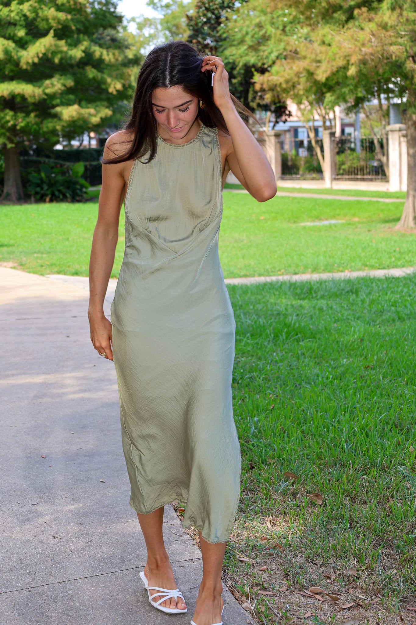A woman wearing a sleeveless olive green midi dress and white strappy sandals walking on a paved path in a park with green grass and trees.