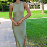 A woman wearing a sleeveless sage green midi dress with a high neckline and side slit, walking outdoors on a paved path next to a grassy park area.
