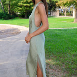 A woman wearing a sage green sleeveless midi dress with a side slit, standing on a concrete path in a park with green grass and trees in the background.