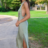 A woman wearing a sage green sleeveless midi dress with a side slit, standing on a concrete path in a park with green grass and trees in the background.