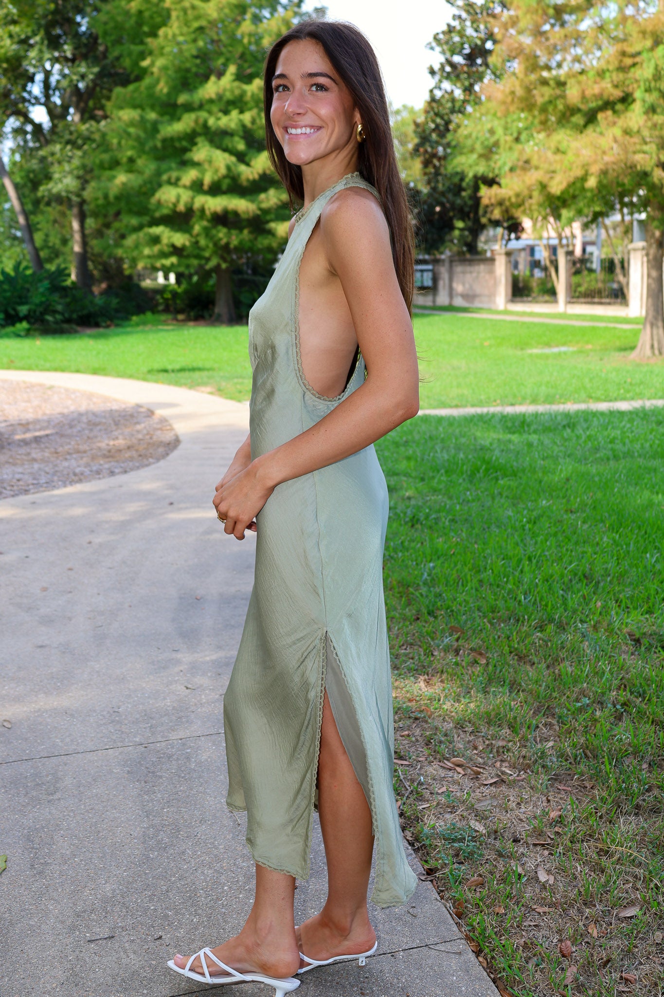 A woman wearing a sage green sleeveless midi dress with a side slit, standing on a concrete path in a park with green grass and trees in the background.