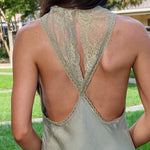 A woman shown from behind wearing a light sage green satin slip dress with a lace-trimmed open back, set against a blurred outdoor background of green grass and trees.