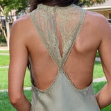 A woman shown from behind wearing a light sage green satin slip dress with a lace-trimmed open back, set against a blurred outdoor background of green grass and trees.