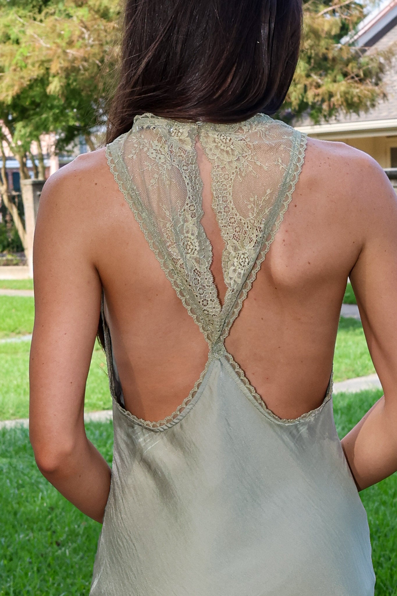 A woman shown from behind wearing a light sage green satin slip dress with a lace-trimmed open back, set against a blurred outdoor background of green grass and trees.