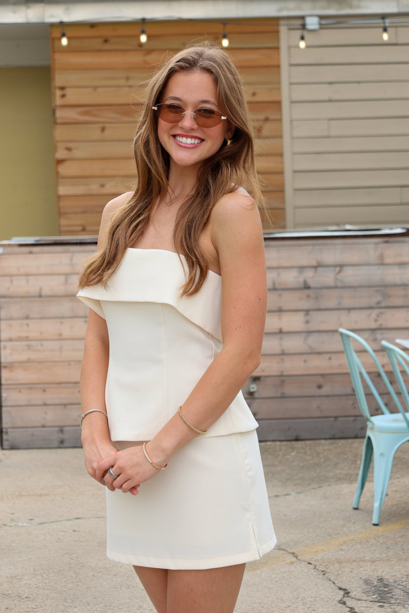 A woman wearing a off white strapless ruffled top and matching mini skirt, styled with sunglasses and bracelets, standing outdoors in front of a wooden wall.