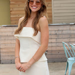A woman wearing a off white strapless ruffled top and matching mini skirt, styled with sunglasses and bracelets, standing outdoors in front of a wooden wall.