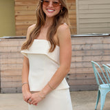 A woman wearing a off white strapless ruffled top and matching mini skirt, styled with sunglasses and bracelets, standing outdoors in front of a wooden wall.