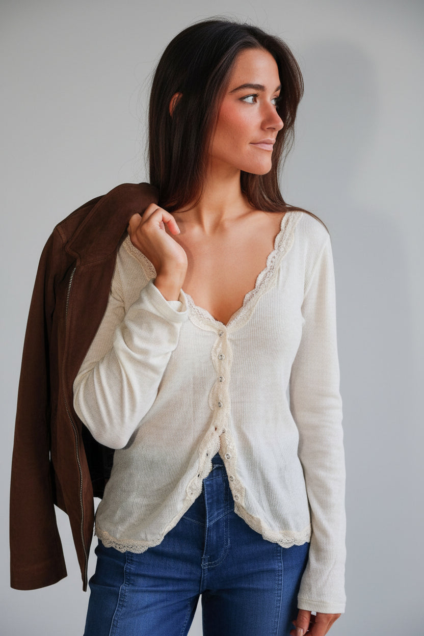 Woman wearing a beige cardigan over a white shirt and blue jeans, leaning against a wall.