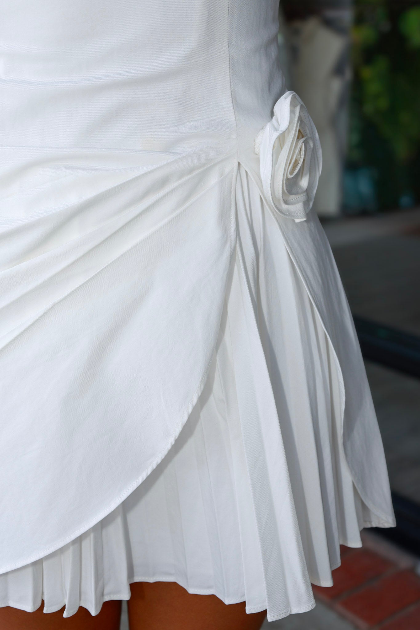Close-up of a white draped skirt with pleated detailing and a side fabric flower embellishment, shown on a model.
