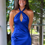 A woman modeling a royal blue satin halter mini dress with a keyhole cutout, standing on a wooden deck porch with trees in the background.