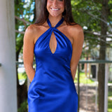 A woman modeling a royal blue satin halter mini dress with a keyhole cutout, standing on a wooden deck porch with trees in the background.