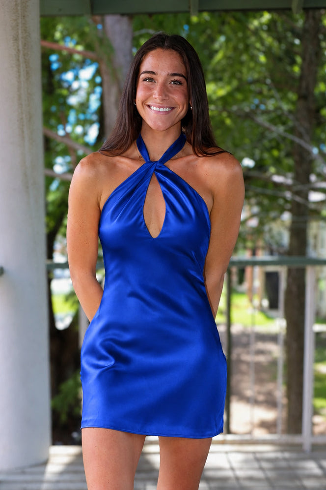 A woman modeling a royal blue satin halter mini dress with a keyhole cutout, standing on a wooden deck porch with trees in the background.