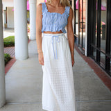 Woman wearing a blue top and white skirt standing on a sidewalk.