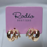 A pair of gold hoop earrings with twisted rope details and small clear crystal accents, displayed on a pink Rodéo Boutique card against a light gray background.