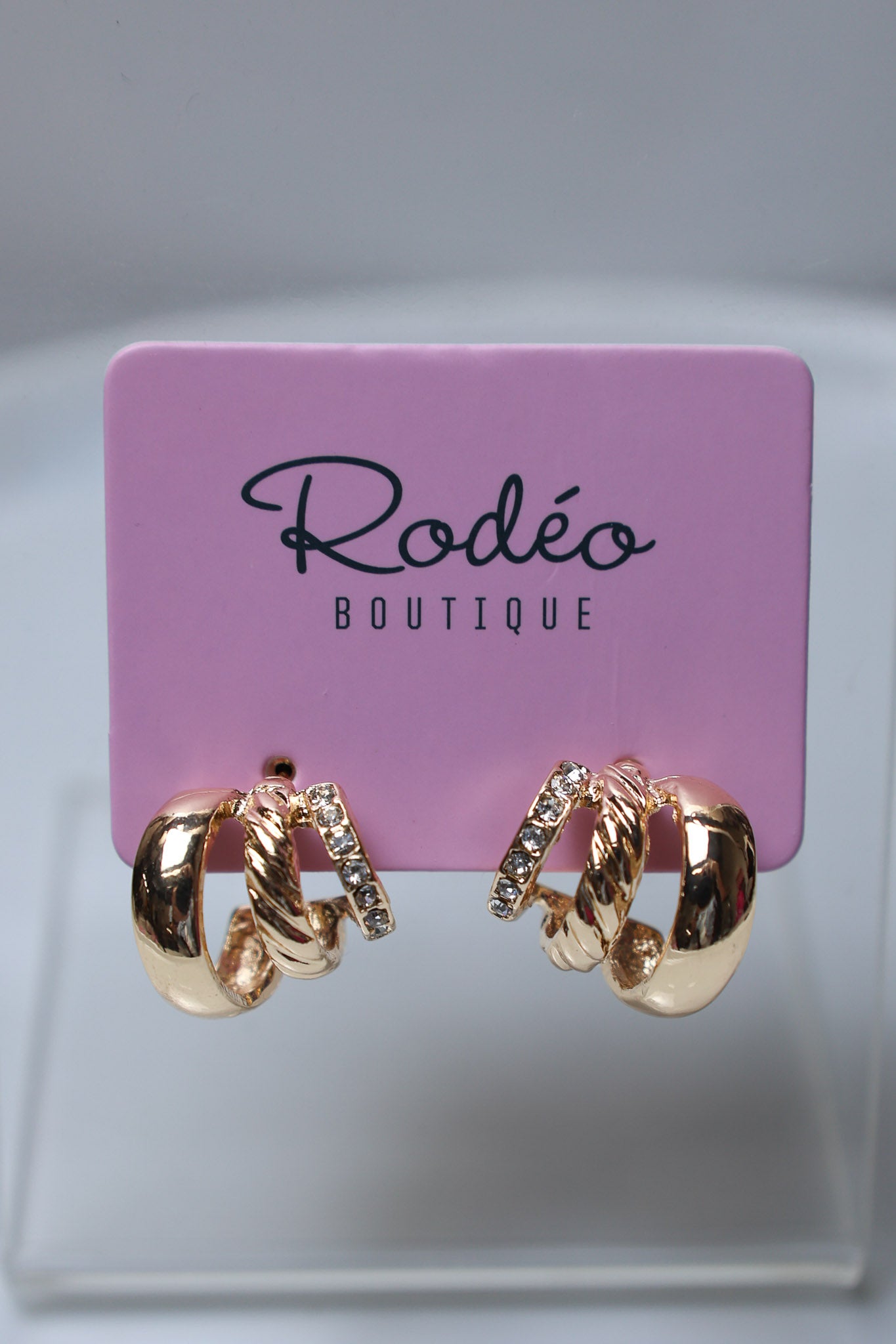 A pair of gold hoop earrings with twisted rope details and small clear crystal accents, displayed on a pink Rodéo Boutique card against a light gray background.