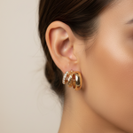 A close-up side profile of a woman's ear wearing a pair of gold hoop earrings with small clear stones, against a soft beige background.