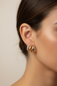 A close-up side profile of a woman's ear wearing a pair of gold hoop earrings with small clear stones, against a soft beige background.