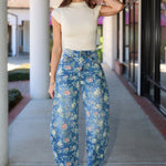 A woman stands on a covered walkway wearing a cream short-sleeve mock-neck top and blue floral barrel jeans with white heeled sandals.