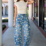 A woman stands on a covered walkway wearing a cream short-sleeve mock-neck top and blue floral barrel jeans with white heeled sandals.