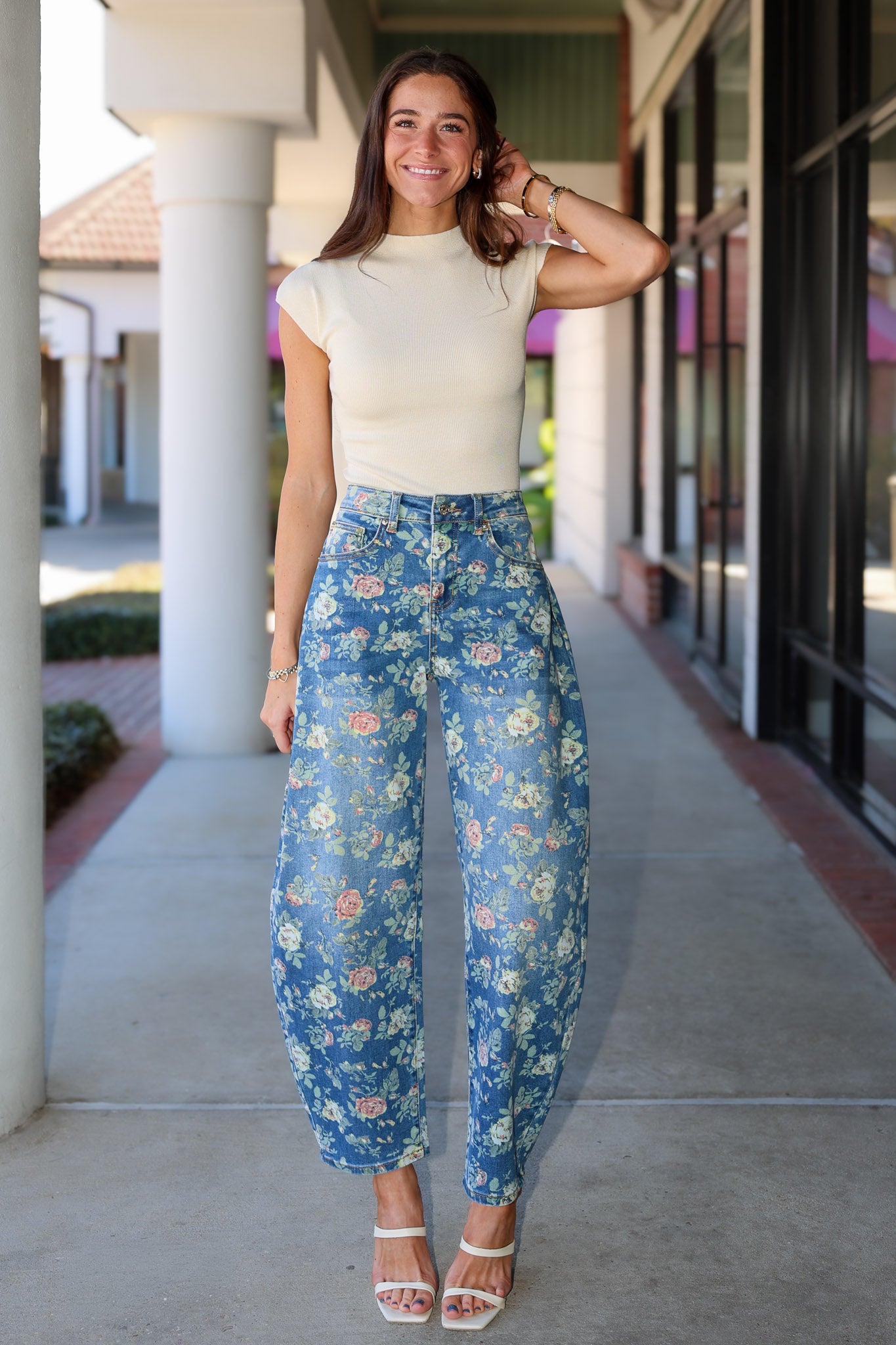 A woman stands on a covered walkway wearing a cream short-sleeve mock-neck top and blue floral barrel jeans with white heeled sandals.