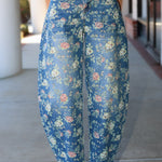 A person wearing high-waisted, wide-leg cropped pants with a blue floral print, paired with a light beige top and white heeled sandals, standing on a concrete walkway.