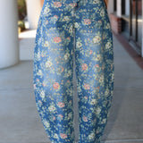 A person wearing high-waisted, wide-leg cropped pants with a blue floral print, paired with a light beige top and white heeled sandals, standing on a concrete walkway.