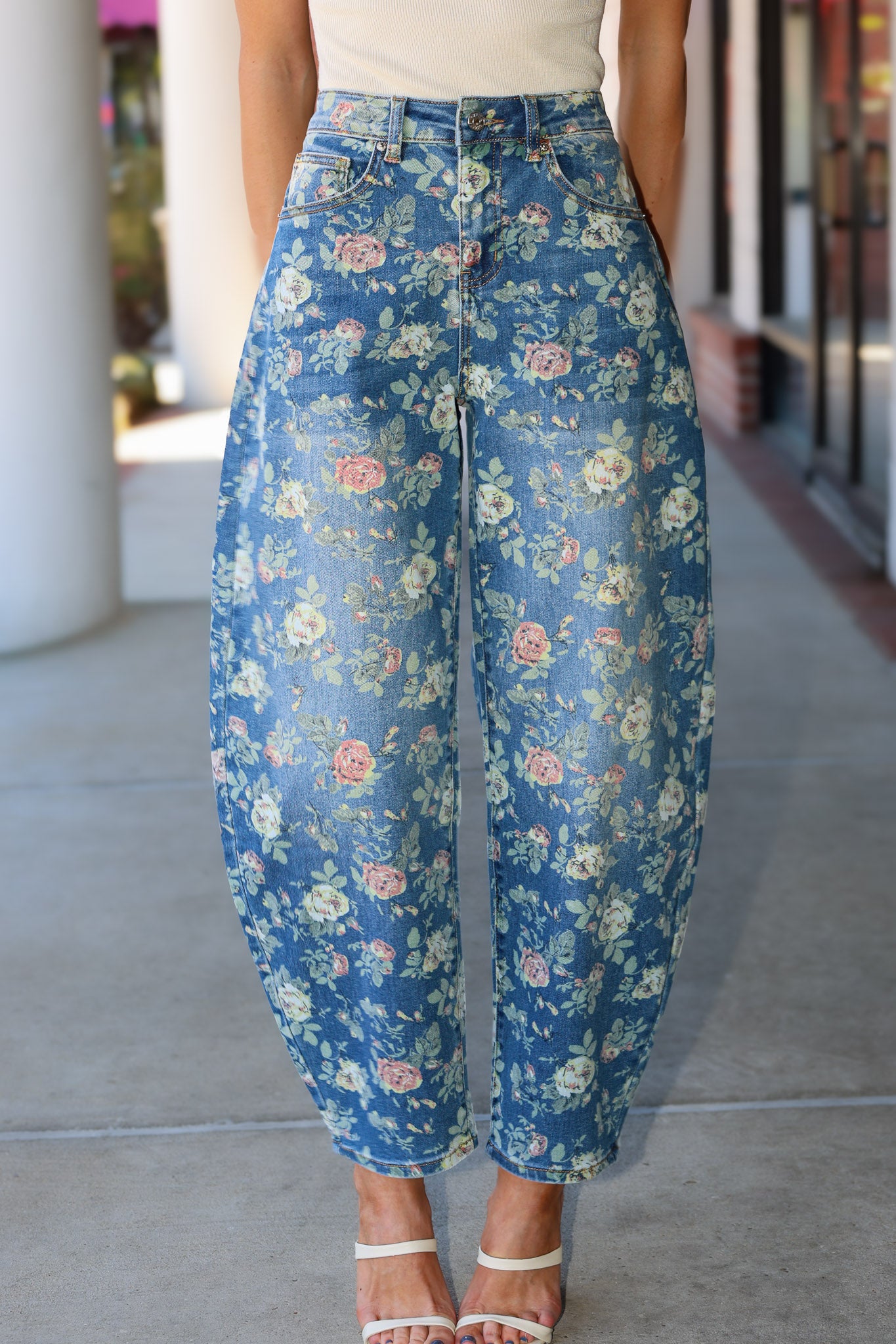 A person wearing high-waisted, wide-leg cropped pants with a blue floral print, paired with a light beige top and white heeled sandals, standing on a concrete walkway.