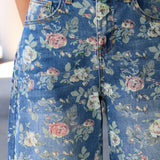 Close-up of a person wearing blue floral jeans with a pink and green rose print, paired with a light beige top.