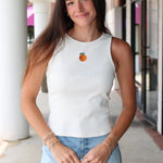 Woman wearing Celestine Tank a white sleeveless shirt with embroidered orange and denim shorts outdoors.