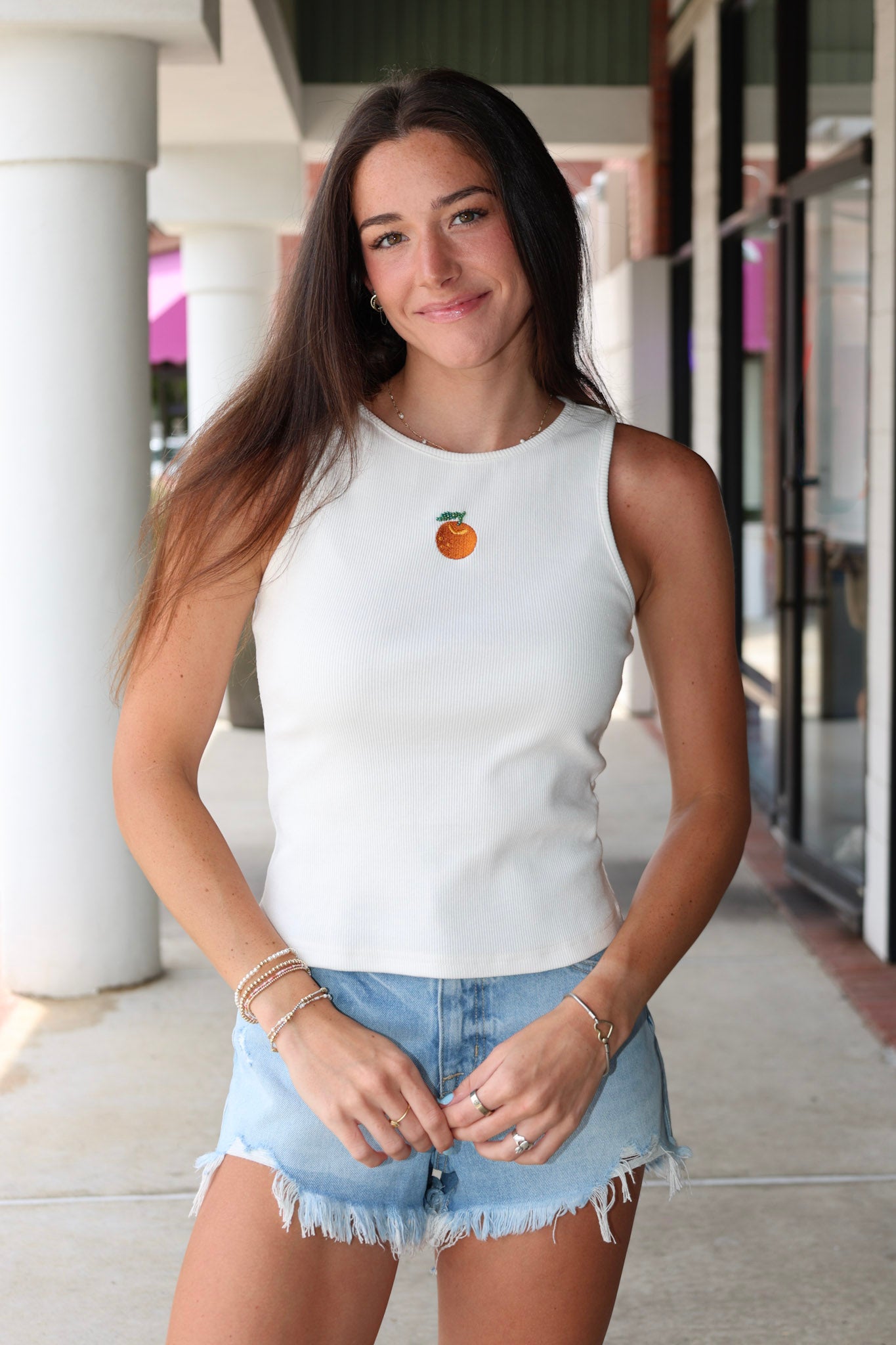 Woman wearing Celestine Tank a white sleeveless shirt with embroidered orange and denim shorts outdoors.