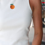 Woman wearing Celestine Tank a white sleeveless shirt with embroidered orange and denim shorts outdoors.