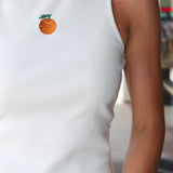 Woman wearing Celestine Tank a white sleeveless shirt with embroidered orange and denim shorts outdoors.