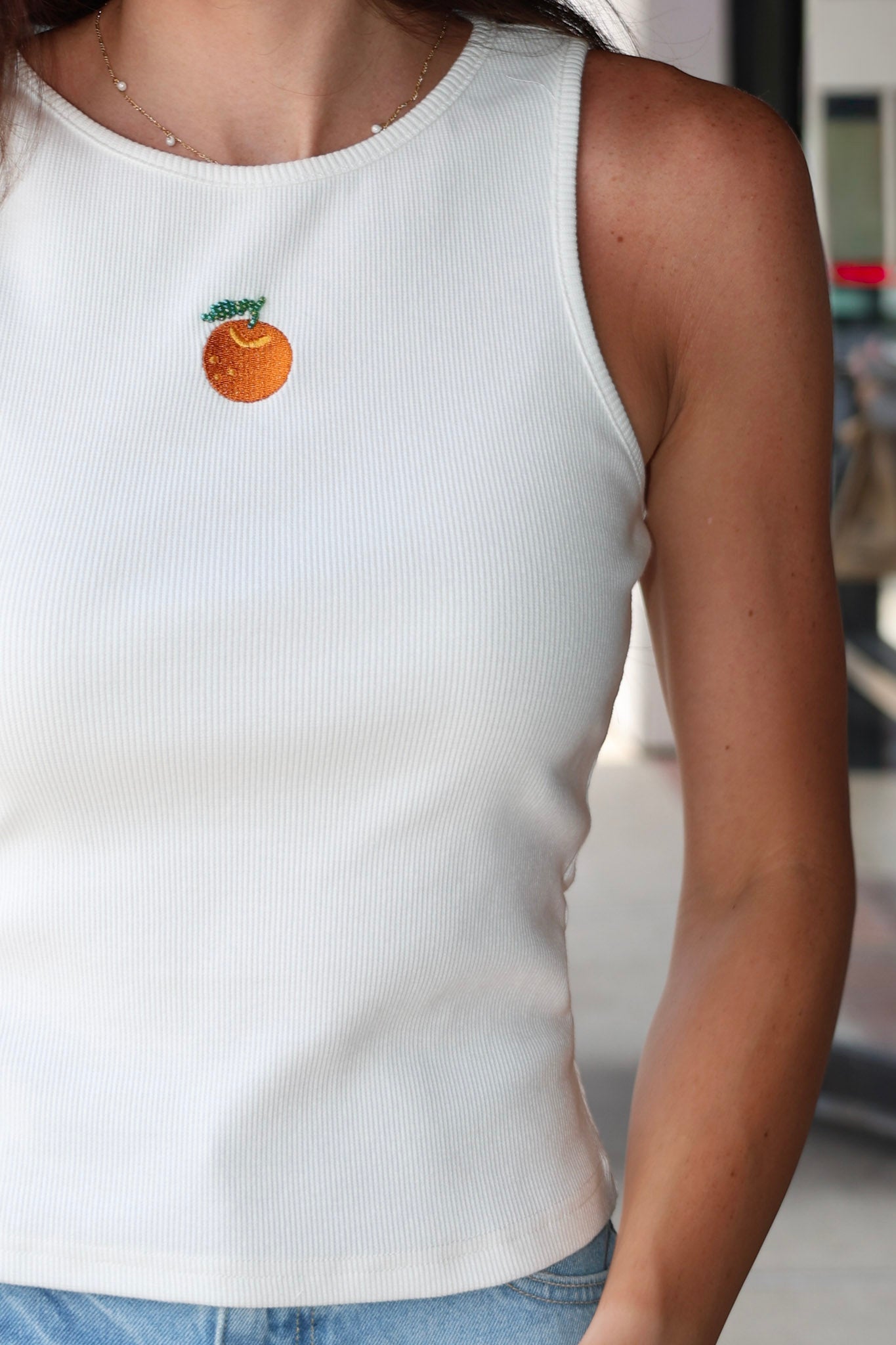Woman wearing Celestine Tank a white sleeveless shirt with embroidered orange and denim shorts outdoors.