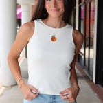 Woman wearing Celestine Tank a white sleeveless shirt with embroidered orange and denim shorts outdoors.
