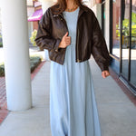 A woman wearing a light blue Charlotte midi dress by ZSupply with a dark brown leather jacket, and brown leather boots for a fall look.