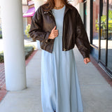 A woman wearing a light blue Charlotte midi dress by ZSupply with a dark brown leather jacket, and brown leather boots for a fall look.