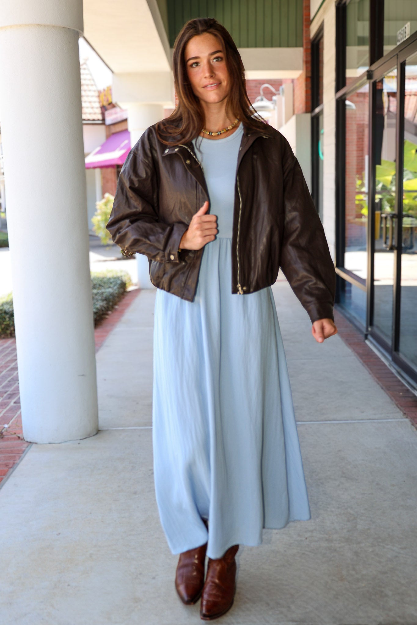 A woman wearing a light blue Charlotte midi dress by ZSupply with a dark brown leather jacket, and brown leather boots for a fall look.