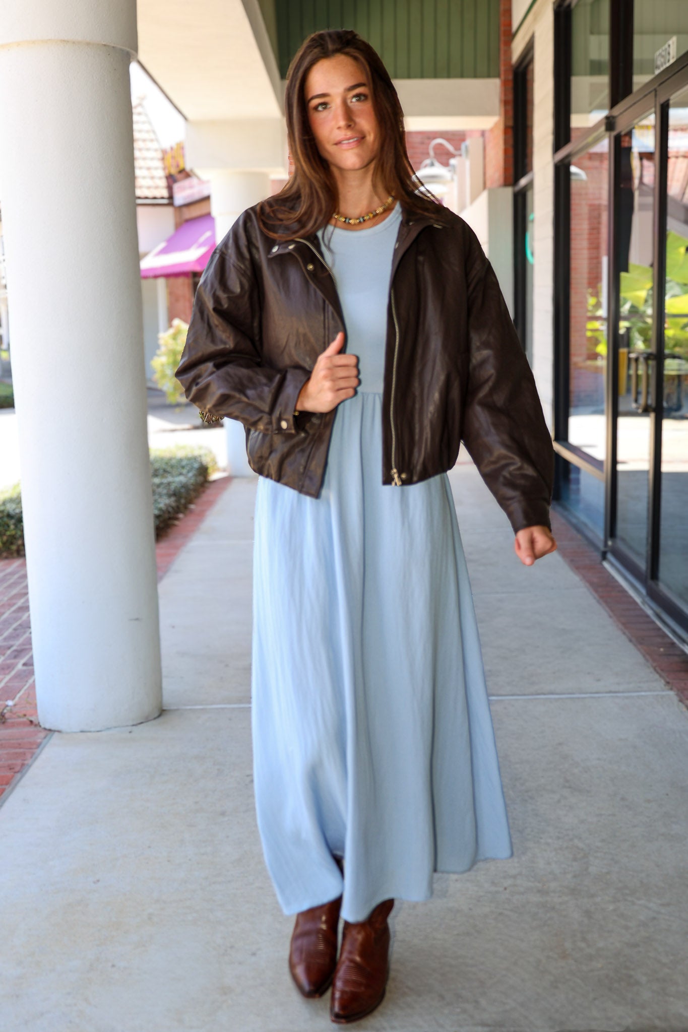 A woman wearing a light blue Charlotte midi dress by ZSupply with a dark brown leather jacket, and brown leather boots for a fall look.