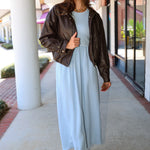 A woman wearing a light blue Charlotte midi dress by ZSupply with a dark brown leather jacket, and brown leather boots for a fall look.