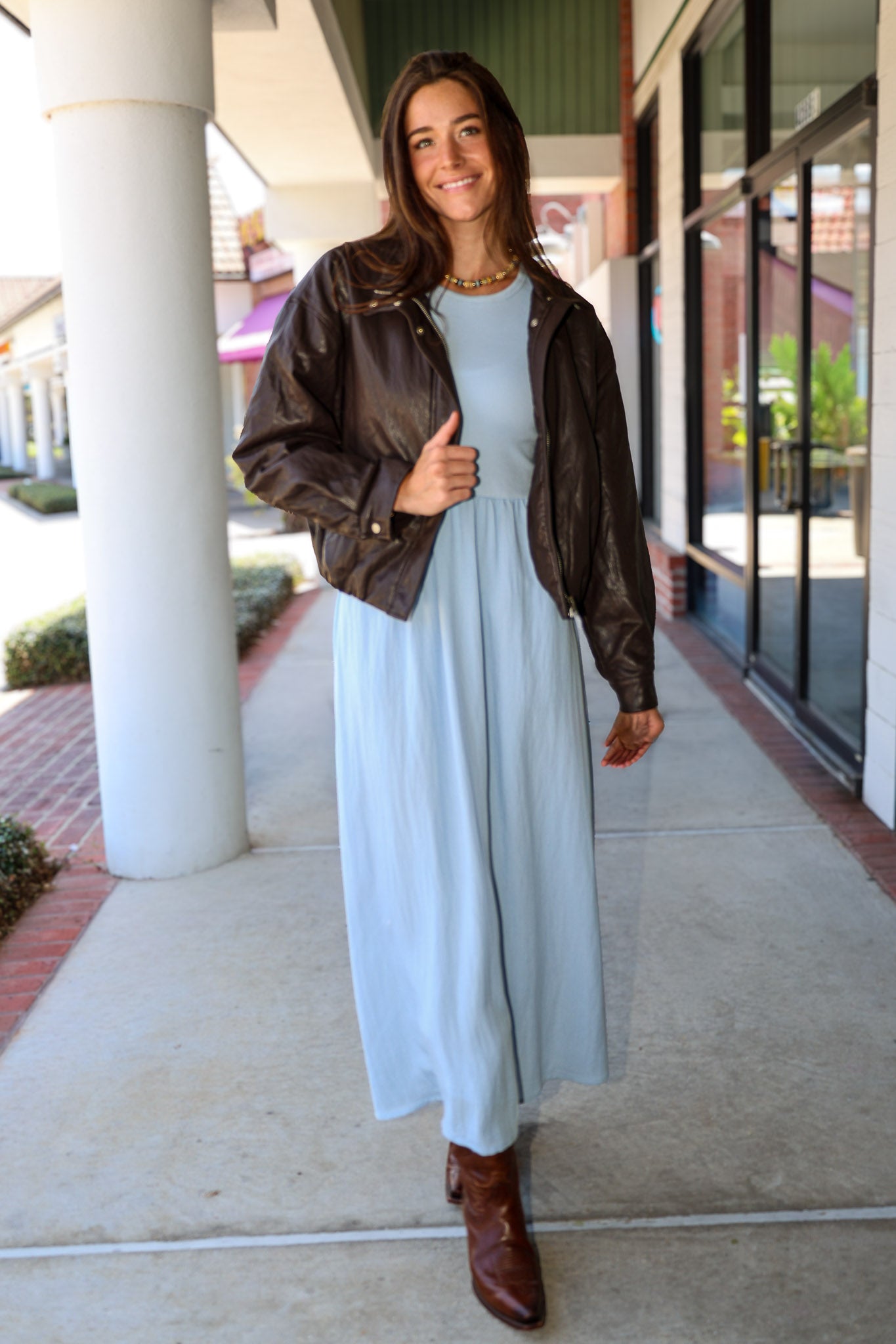 A woman wearing a light blue Charlotte midi dress by ZSupply with a dark brown leather jacket, and brown leather boots for a fall look.